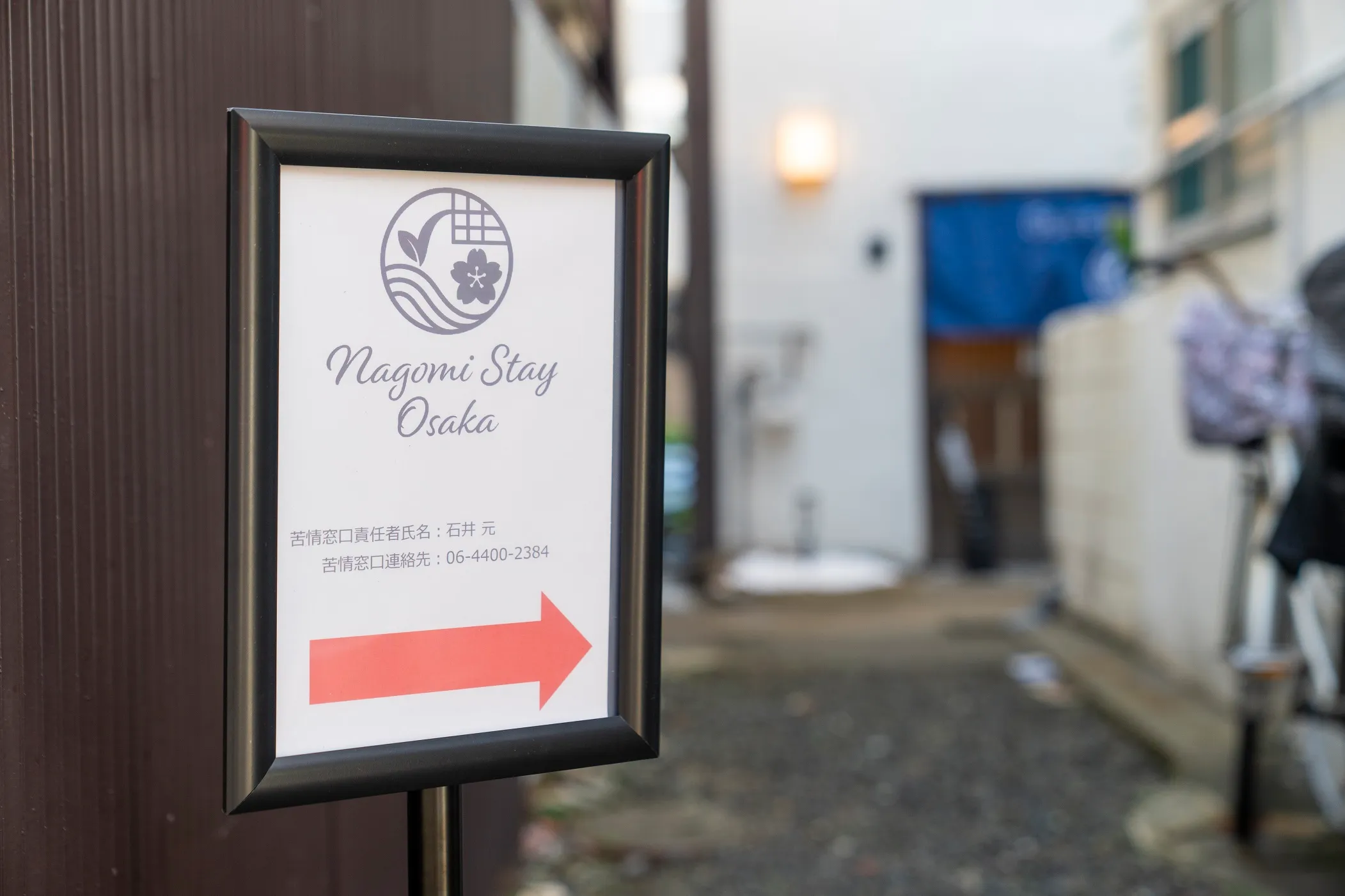 A framed sign on the street reading Nagomi Stay Osaka, with an arrow pointing down the lane towards the entrance